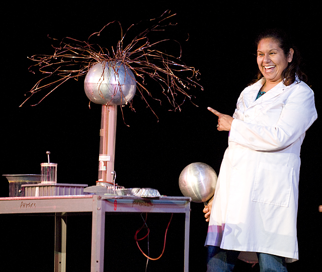Maria Vaughn in a lab coat pointing at a Van de Graaff generator