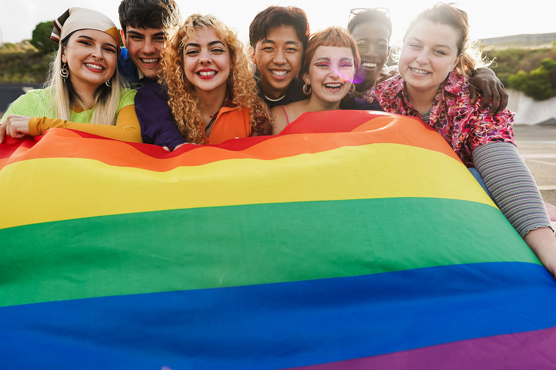 A collection of young people are all smiles around a rainbow flag.