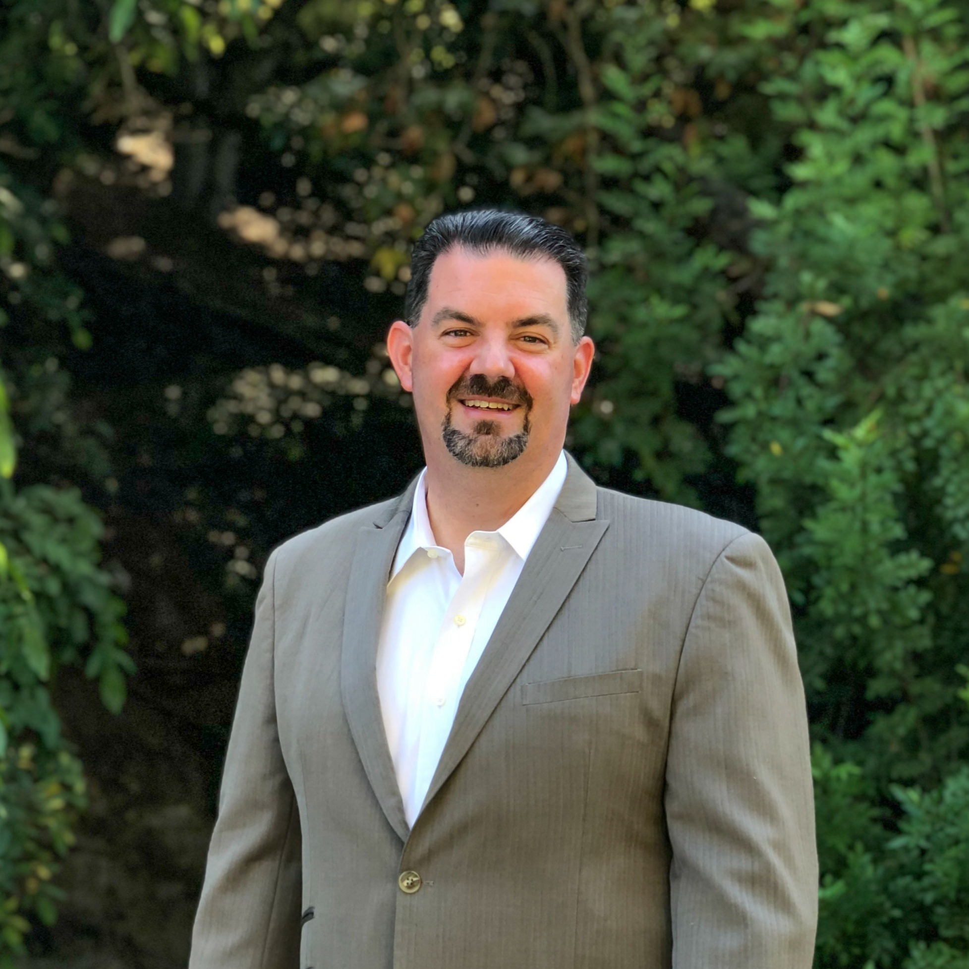 Portrait of white man with dark short hair and a well-trimmed beard in front of a hedge.