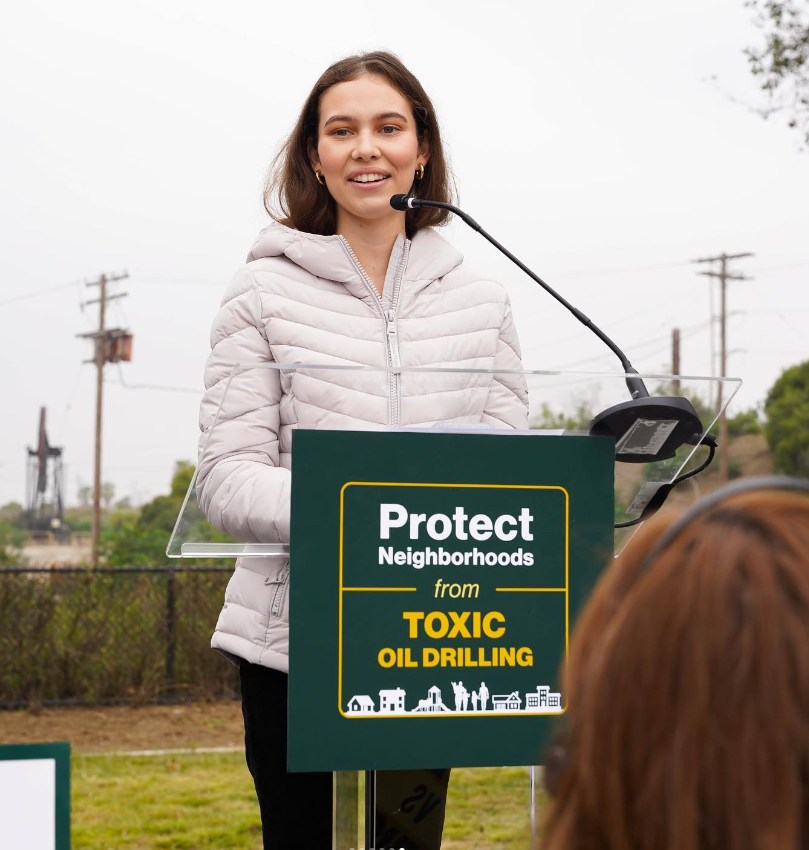 A dark haired young woman at podium advocating for urban drilling to stop
