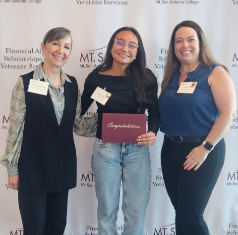 Three women stand close together smiling at scholarship ceremony.