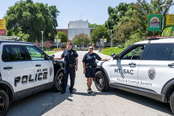 A female police chief poses with a black and white car and another officer stands to her left next to a white car