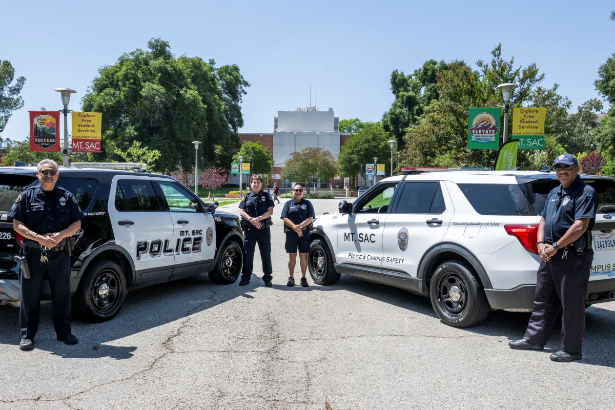 A male and female police officer stand next to a black and white car and to their left is a female and male officer standing next to a white car.