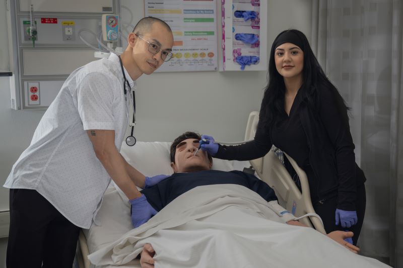 A male nursing student with a stethoscope leans over a simulator that looks like a man and a female student with long black hair inserts a thermometer into the mannequin's mouth. used to