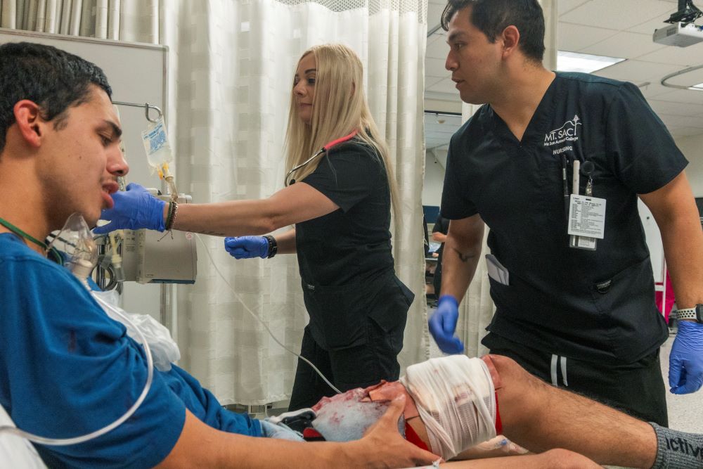 A male student with a realistic leg wound, complete with blood, is tended to by a blonde female nursing student while a male Filipino nurse supervises the situation.