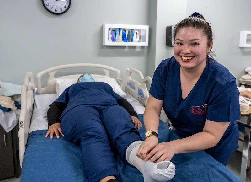 A Latina woman dressed in a Mt. SAC dark blue Nursing Assistant uniform smiles at the camera while simulating working on a patient's foot.
