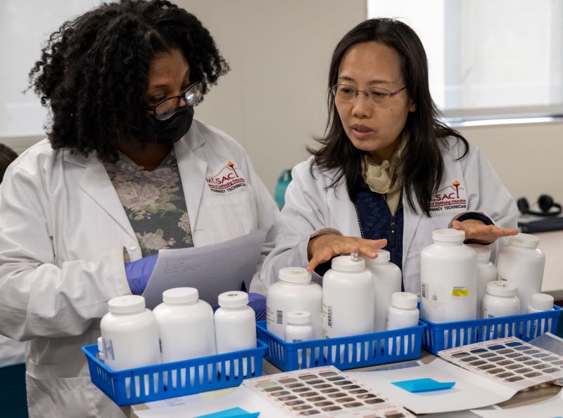 A black woman and an Asian woman look at pill bottles as part of the Pharmacy Tech program.