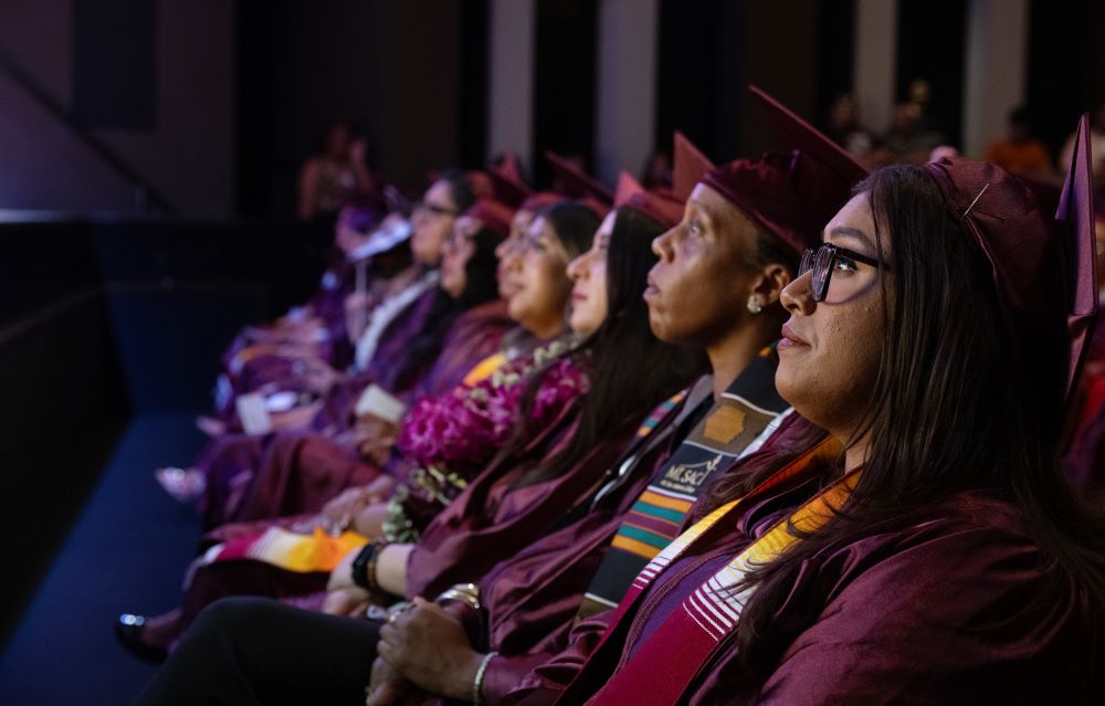 Women dressed in maroon graduation regalia in profile look ahead as they listen to a speaker in front of them.