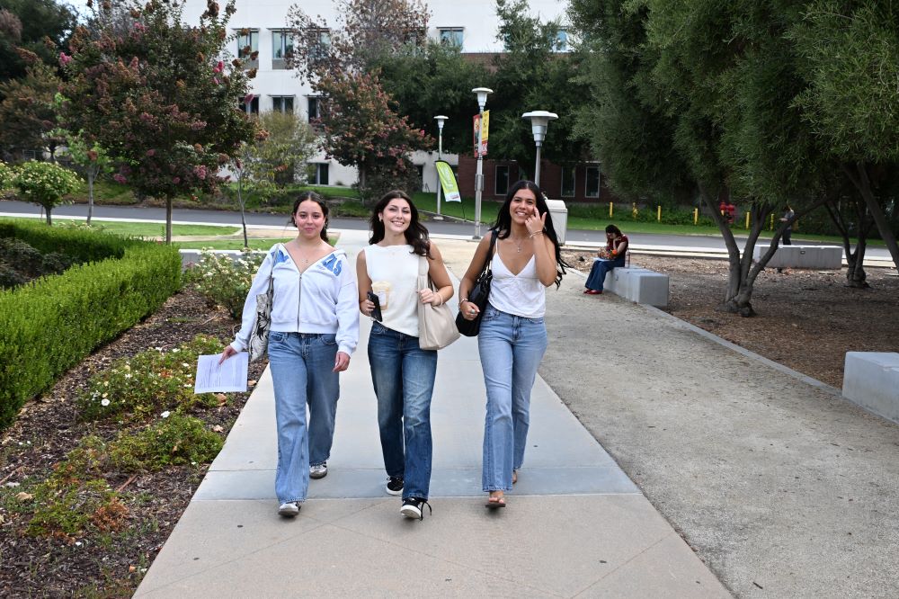 Three women in their early 20s wearing jeans smile as they walk toward the camera.