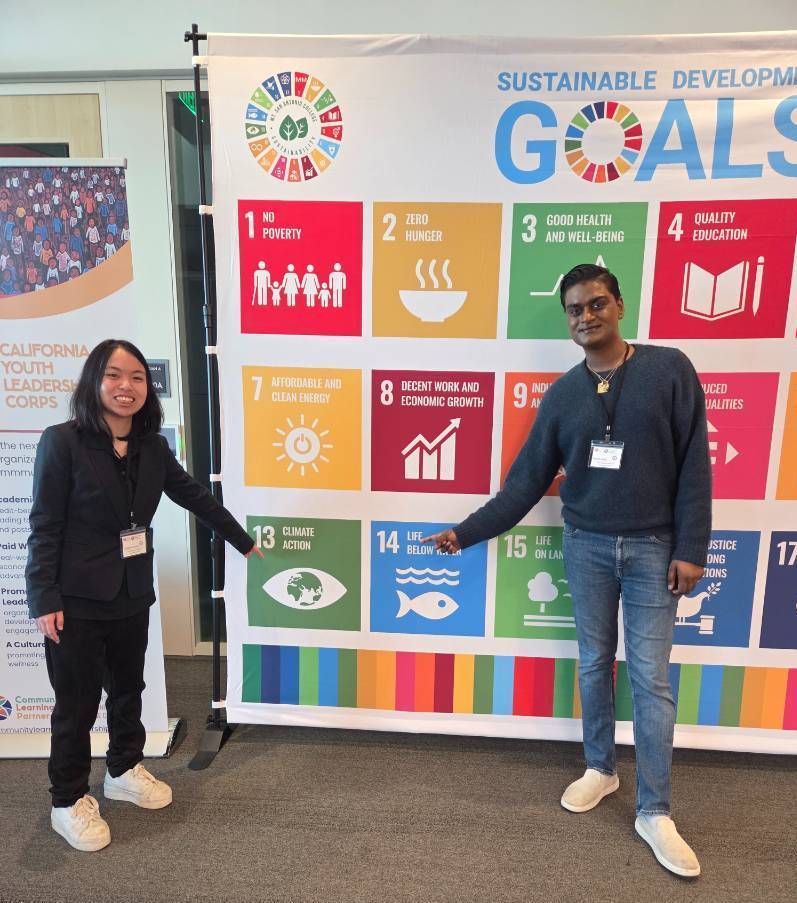 A female student and a male student stand in front of a large banner that lists all of the areas of sustainability Mt SAC focuses on