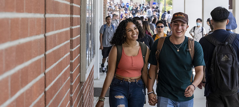 A student couple hold hands walking with line for student IDs in the background