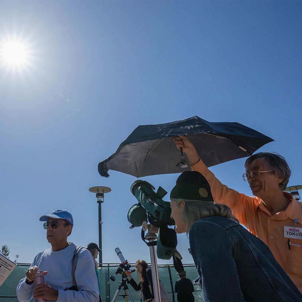 person looking through a telescope while another hold umbrella to shade the view